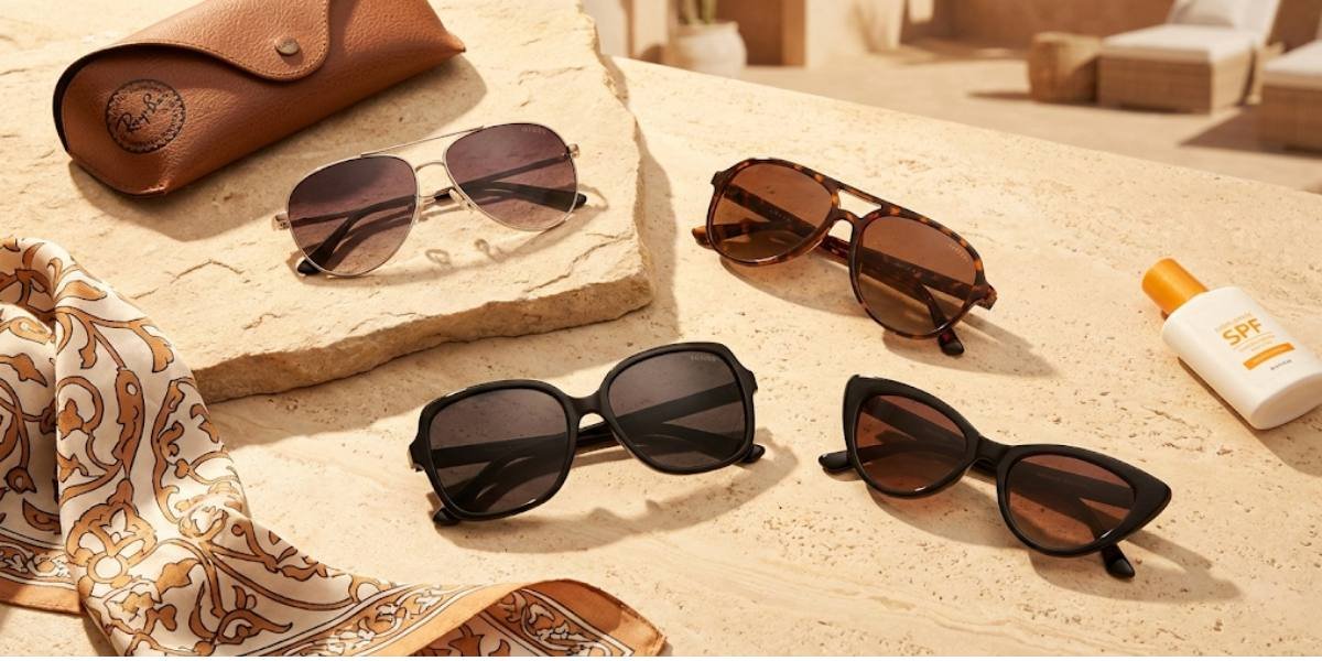 A flat lay collection of trending women's sunglasses for 2025, featuring aviator, oversized, and cat-eye styles arranged on a sunlit stone surface alongside a leather case, silk scarf, and sunscreen.
