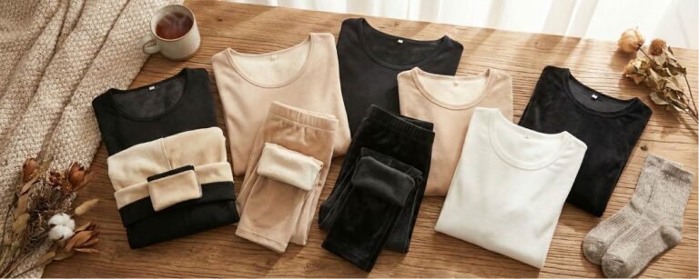 عرض مسطح لمجموعة متنوعة من أفضل ملابس داخلية حرارية للنساء، تشمل أطقم مبطنة بالصوف وأخرى رقيقة بألوان الأسود والبيج والأبيض. الصورة تعكس أجواء شتوية دافئة ومريحة على طاولة خشبية مع كوب شاي وبطانية.